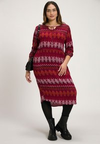 Rotes gemustertes Kleid mit geometrischen Designs, dreiviertelÄrmel und einem Bindekragen; kombiniert mit schwarzen Leggings und klobigen schwarzen Stiefeln.