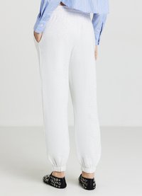 Un pantalon de jogging blanc en matériau doux, avec des poignets élastiqués et des poches latérales, associé à une chemise rayée bleue et des chaussures noires ornées.