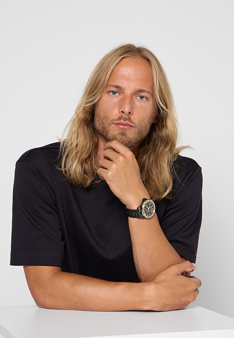 Montre noire avec un cadran rond, accents dorés et bracelet noir. Le modèle porte un t-shirt noir, posé avec les bras croisés contre un fond clair.