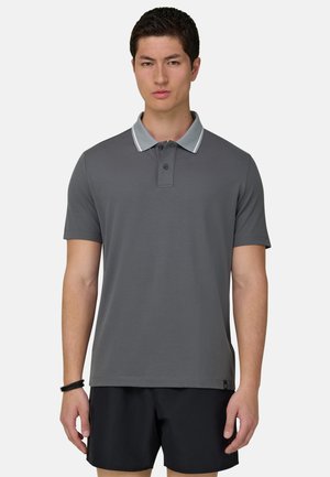 Jonge man die een grijs poloshirt met korte mouwen en een lichtgrijze kraag en zwarte korte broek draagt, staand tegen een effen achtergrond.