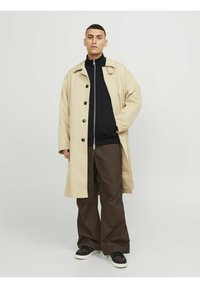 Beige yderjakke med knap-front design, båret over en sort lynlås hoodie og brune brede bukser, parret med sorte sneakers.