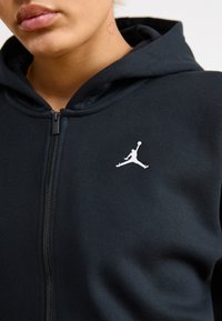 Sudadera con cremallera negra que presenta un logo Jumpman blanco, puños y bajo acanalados, y una textura suave. Detalle de cremallera visible en la parte frontal.