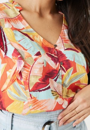 Femme portant un chemisier fleuri coloré avec col en V et boutons, associé à un jean taille haute bleu clair et une boucle de ceinture noire.