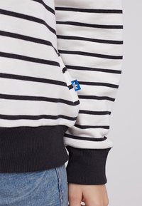 Pruhovaná mikina, bílá s černými pruhy, s námořnickým žebrovaným spodním lemem a manžetami. Malá modrá označovací nálepka na boku. Měkký bavlněný materiál.