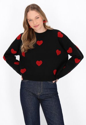 Zwarte trui met een cropped ontwerp, voorzien van gestructureerde rode hartpatronen. Geselecteerd bij donkere spijkerbroek, tegen een witte achtergrond.