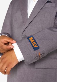 Ręka dostosowująca biały mankiet pod szarym rękawem marynarki z pomarańczową i niebieską naszywką "DAILY" oraz trzema szarymi guzikami.