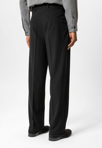 Zwarte op maat gemaakte broek met een rechte pasvorm, gladde stof en achterzakken. Draagt een lichtgrijs overhemd met knopen en zwarte schoenen.