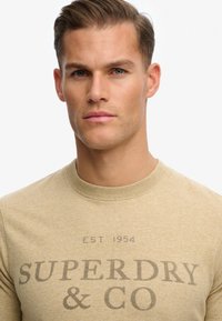 Sudadera de algodón beige con cuello redondo, que presenta un gran logo impreso "SUPERDRY & CO" en gris oscuro y un acabado suave y texturizado.