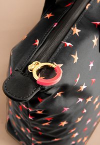 Juoda audinio rankinė su raudonomis ir oranžinėmis žvaigždžių raštais, turinti auksinį užtrauktuko traukimą su rožinės gumos laikymo žiedu.