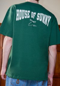 Άτομο που φοράει πράσινη μπλούζα με λευκή επιγραφή "HOUSE OF SUNNY" και μικρότερο κείμενο "Hackney London" στο πίσω μέρος, συνδυασμένη με μπλε τζιν.