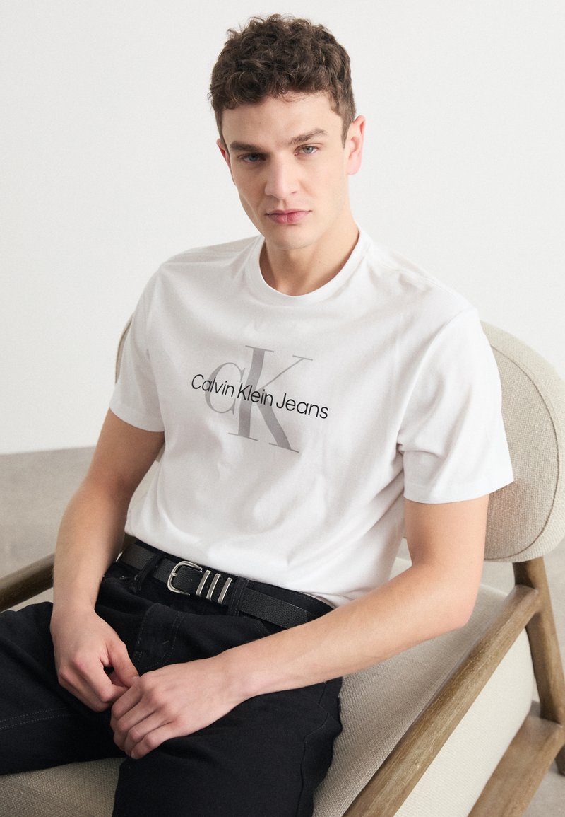 T-shirt bianco in cotone con logo "Calvin Klein Jeans" grigio, indossato con pantaloni neri e una cintura nera strutturata, seduto su una sedia.