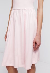 Femme portant une robe texturée rose clair, longueur genou, avec une taille cintrée et une jupe froncée, se tenant les mains détendues sur les côtés.