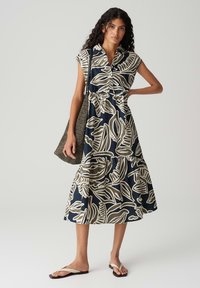 Blaues blumengemustertes Kleid mit kurzen Ärmeln, Kragen und gestuften Saum. Hergestellt aus leichtem Material, kombiniert mit einer strukturierten Tasche und Sandalen.