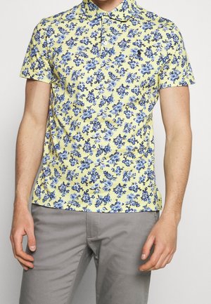 Homme portant un polo jaune à manches courtes avec un motif floral bleu et un pantalon gris, debout les bras détendus le long du corps.