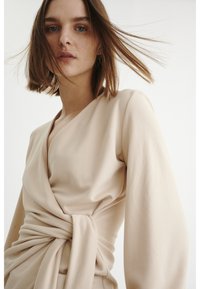 Vestido envolvente bege com um decote em V profundo, mangas longas e um detalhe de nó torcido na cintura. Tecido suave e caimento fluido.