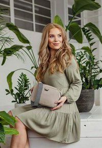 Beige křížová taška s vzorovaným popruhem, s logem. Hladký materiál, obdélníkový tvar a jemné akcenty. Spárovaná s dlouhým zeleným šaty.