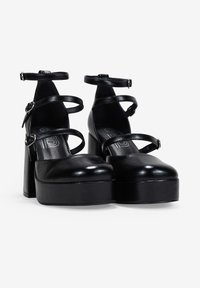 Tacones de plataforma negros con punta redonda, textura de cuero brillante y múltiples correas en el tobillo con hebillas ajustables.