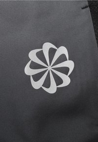 Černá látka s námořnickomodrým logem ve tvaru květu se zakřivenými okvětními lístky, umístěným ve středu na texturovaném povrchu. Žádné další detaily nejsou viditelné.