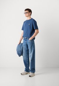 Blå bomulds t-shirt med lille trekantet logo, parret med lyseblå straight-leg jeans og grå sneakers. Holder en denimjakke.