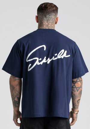 Mann mit Hals- und Armtätowierungen, der ein marineblaues T-Shirt mit großem weißem Schriftzug "Siksilk" auf dem Rücken trägt.