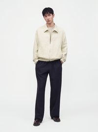Jeune homme portant une veste beige à fermeture éclair, un pantalon sombre et ample, et des chaussures foncées, debout les mains dans les poches devant un fond uni.