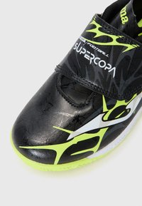 Scarpa da calcio nera con finitura lucida, motivo testurizzato, accenti gialli e una fascetta in Velcro con il marchio "SUPERCOPA".