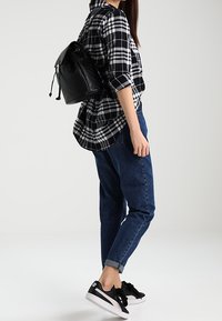 Sac à dos matelassé noir avec fermeture à cordon, porté sur une chemise à carreaux noir et blanc, associé à un jean en denim bleu et des baskets noires.