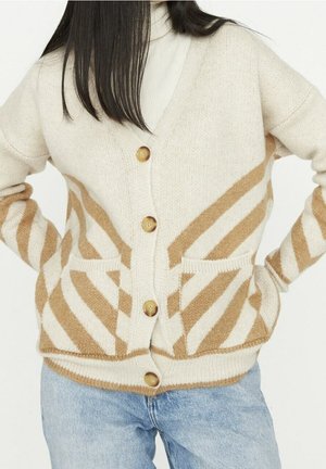 Frau trägt eine beige und braun gestreifte, geknöpfte Strickjacke mit Fronttaschen über hellblauer Jeans, posiert mit den Händen in den Hüften.