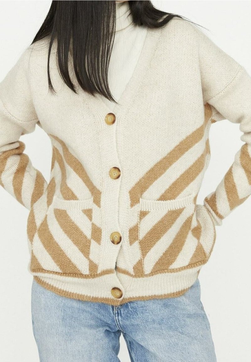Femme portant un cardigan beige et marron à rayures, boutonné avec des poches avant, sur un jean bleu clair, posant les mains sur les hanches.