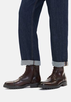 Brązowe skórzane buty z zaokrąglonym noskiem, wiązane na sznurowadła, z teksturowaną podeszwą. Dopasowane do podwiniętych ciemnych dżinsów z jasnoniebieskimi mankietami.
