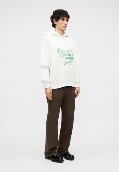 Νεαρός άνδρας με σγουρά μαλλιά, φορεμένος με λευκή μπλούζα με κουκούλα που έχει πράσινη εκτύπωση Döner House, καφέ παντελόνι και μαύρα παπούτσια, στέκεται μπροστά σε απλό φόντο.