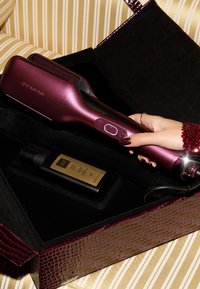 Ghd duet style lisseur en métal bordeaux, au design élégant et avec des commandes par bouton, placé dans un étui doublé en velours noir aux côtés d'un flacon pulvérisateur noir.