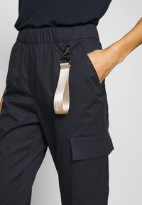Námořnické kalhoty z látky s elastickým pasem, boční kapsou a béžovým popruhem s sponou. Design zahrnuje klopovou kapsu na nohavici.