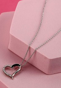 Collana con pendente a forma di cuore in argento, caratterizzato da una superficie lucida con piccoli cristalli trasparenti, sospeso a una delicata catena su uno sfondo rosa.