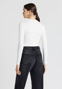 Frau mit dunklen Haaren im Pferdeschwanz, trägt silberne Creolen, ein weißes geripptes Langarmoberteil und schwarze hoch taillierte Jeans, zeigt von hinten.