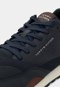 B baskets bleu marine en maille et cuir, avec des lacets foncés et un accent marron. Affiche le logo "TOMMY HILFIGER" sur le côté.