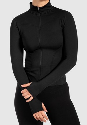 Frau trägt ein eng anliegendes schwarzes langärmliges Zip-up-Oberteil mit Daumenlöchern und steht mit verschränkten Händen vor einem einfarbigen Hintergrund.