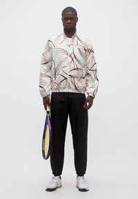 Άνδρας στέκεται κρατώντας ρακέτα τένις, φοράει άσπρο μπουφάν με μαύρες και κόκκινες αφηρημένες γραμμές, μαύρο παντελόνι και άσπρα αθλητικά παπούτσια σε απλό φόντο.