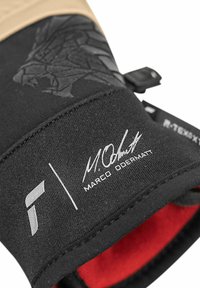 Lähikuva mustastah sportkindadest, millel on tekstuuritud haare, punane sisemine vooder ja "Marco Odermatt" allkiri trükitud randme piirkonda.