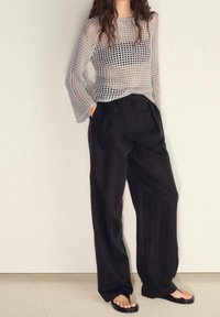 Femme portant un pantalon noir à jambes larges, des tongs noires et un haut à manches longues en maille grise avec une couche noire en dessous, debout les mains dans les poches.