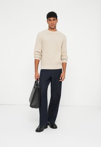 Maglione beige a trama, pantaloni larghi blu navy e scarpe nere. Tiene una borsa tote nera. Design semplice e moderno con colori neutri.