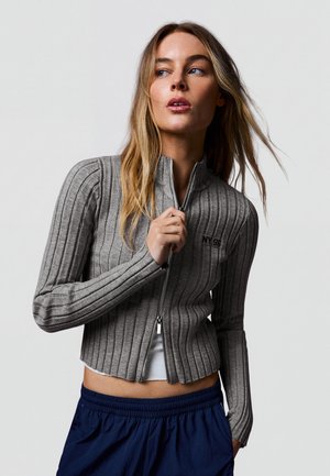 Femme aux longs cheveux blonds portant une veste zippée côtelée grise et un pantalon bleu marine, regardant sur le côté.