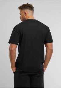 Tricou din bumbac negru, cu croială lejeră, mâneci scurte și guler rotund. Vederea din spate evidențiază cusături curate și o textură netedă.
