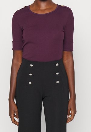 Frau trägt ein lila Kurzarm-Oberteil mit goldenen Knöpfen und schwarze, hoch taillierte Hosen mit dekorativen silbernen Knöpfen an der Vorderseite.