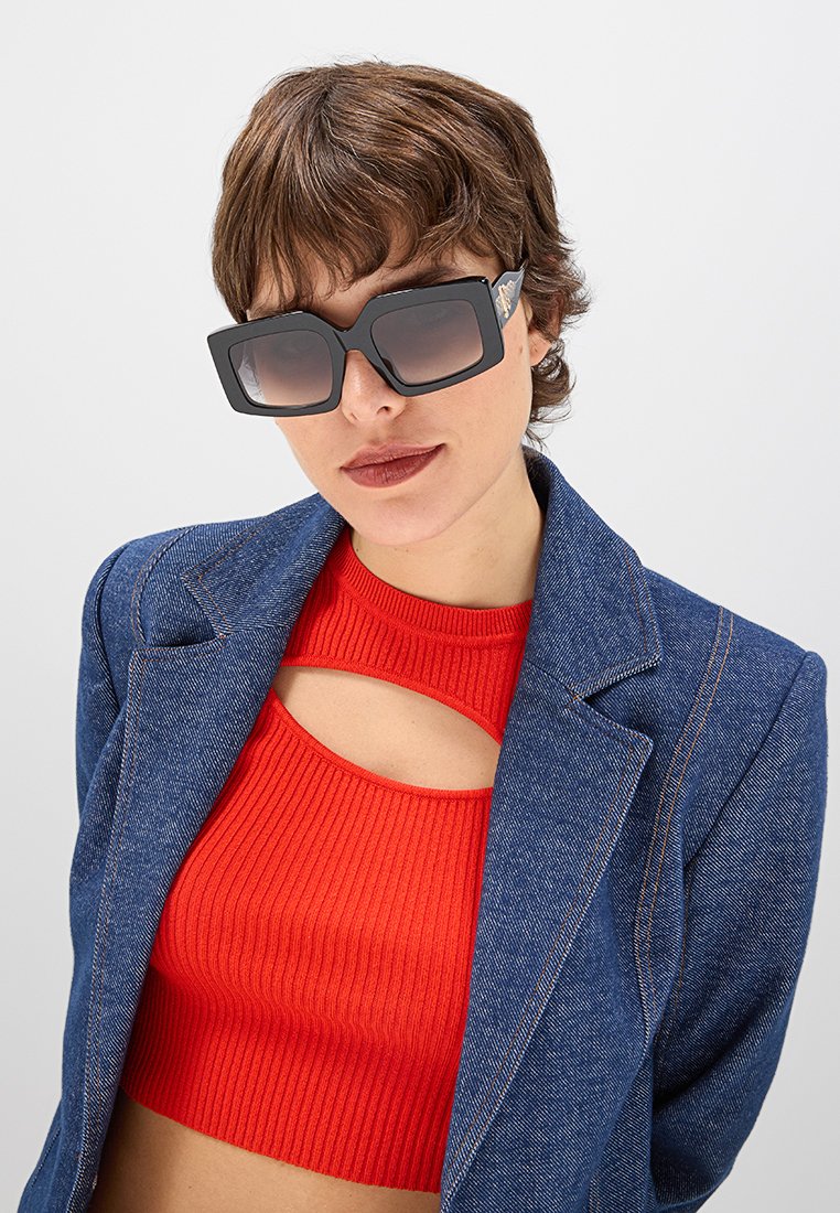 Schwarze quadratische Sonnenbrille mit verlaufenden Gläsern, ausgestattet mit dicken Rahmen und strukturierten Ecken, getragen mit einem roten, gerippten Crop-Top und einer Jeansjacke.