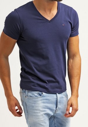 Granatowy t-shirt z dekoltem w serek, wykonany z bawełny, dopasowany krój z krótkimi rękawami. Małe logo na piersi. Połączony z jasnoniebieskimi jeansami.