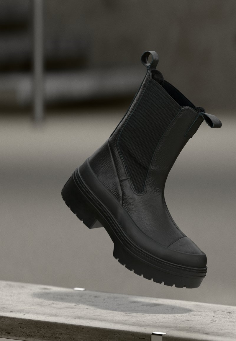 Bottine en cuir noir avec semelle en caoutchouc épaisse et languettes de tirage, présentée suspendue au-dessus d'une surface en béton dans un décor industriel minimaliste.