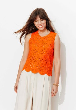 Jeune femme souriante, yeux fermés, portant de grandes boucles d'oreilles en créole, un haut orange en crochet sans manches, un pantalon large plissé blanc et un bracelet en or.