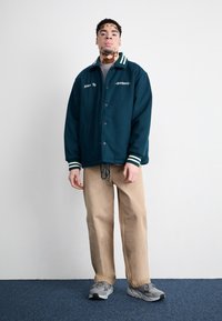 Grønn og hvit stripete kragejakke med knapper, kombinert med beige bukser og grå joggesko. Modellen står mot en ensfarget bakgrunn.