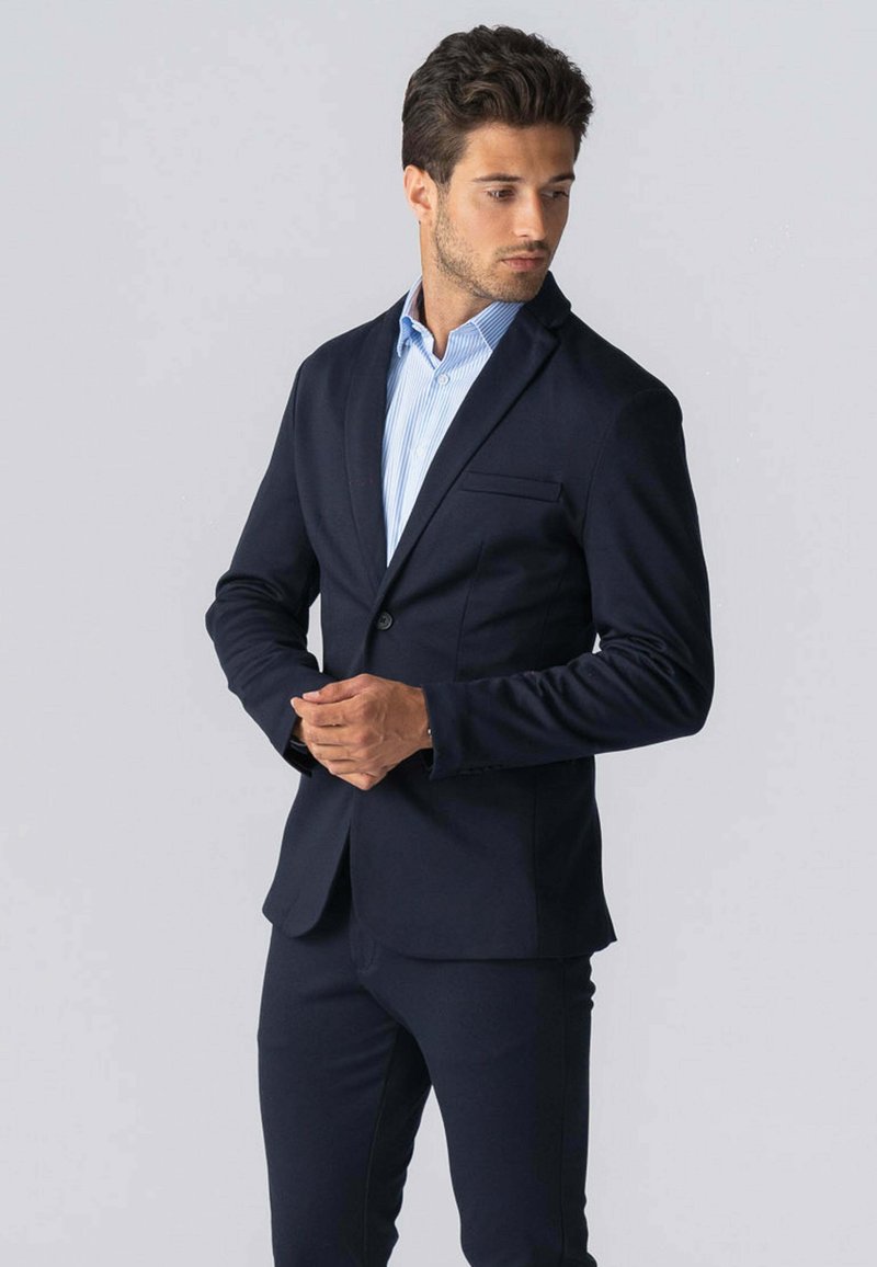 Mørk navy blazer lavet af et glat, tætsiddende stof, med en enkelt knaplukning, klapkrave og forlommer. Stylet med en lysblå stribet skjorte.